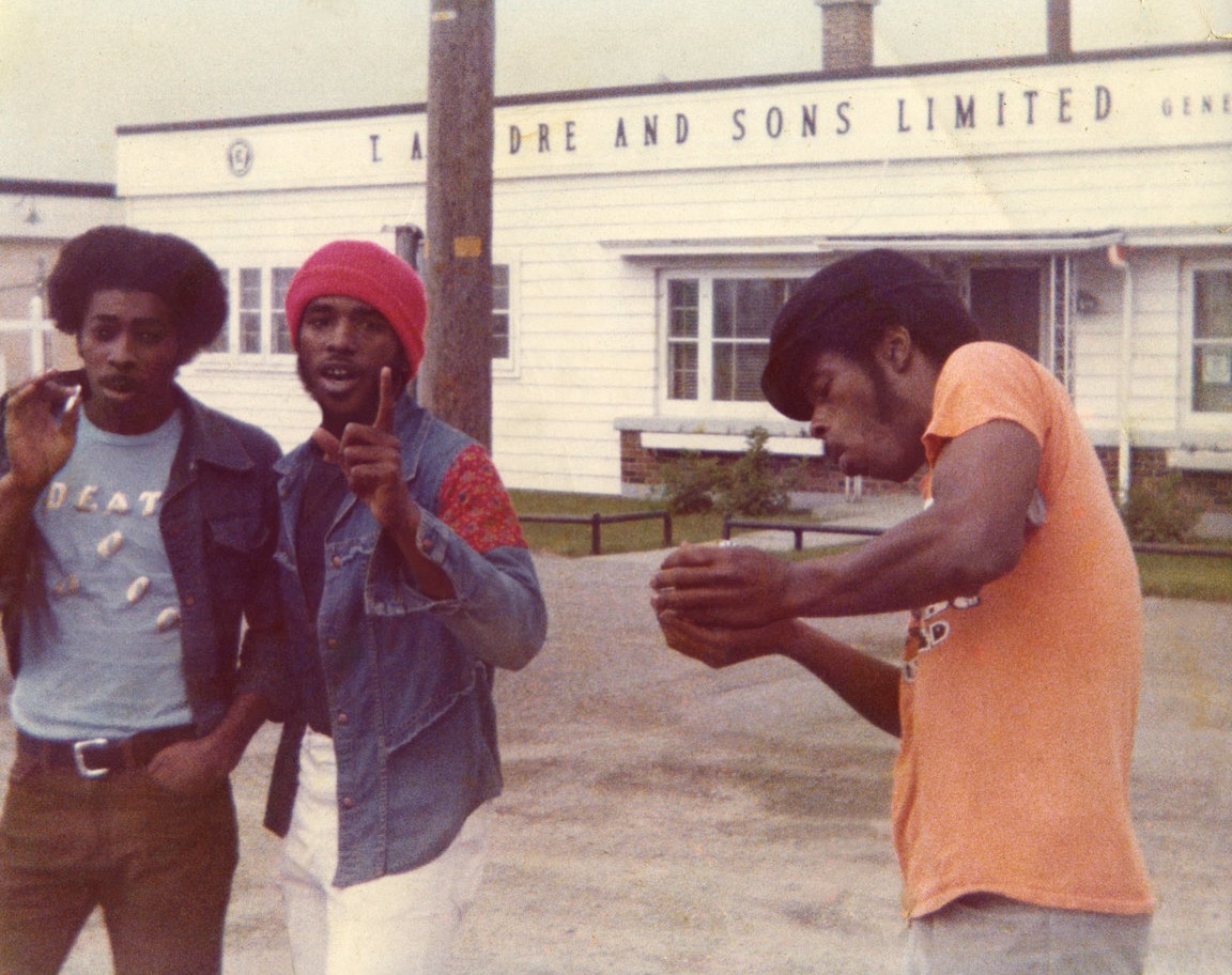 Three young men on a paved forecourt outside a white clapboard building lettered TAYLORE AND SONS LIMITED. The man on the left wears a t-shirt reading DEATH; the man in the centre wears a red knitted cap and points at the camera; the man on the right, in an orange t-shirt, looks down at his cupped hands.