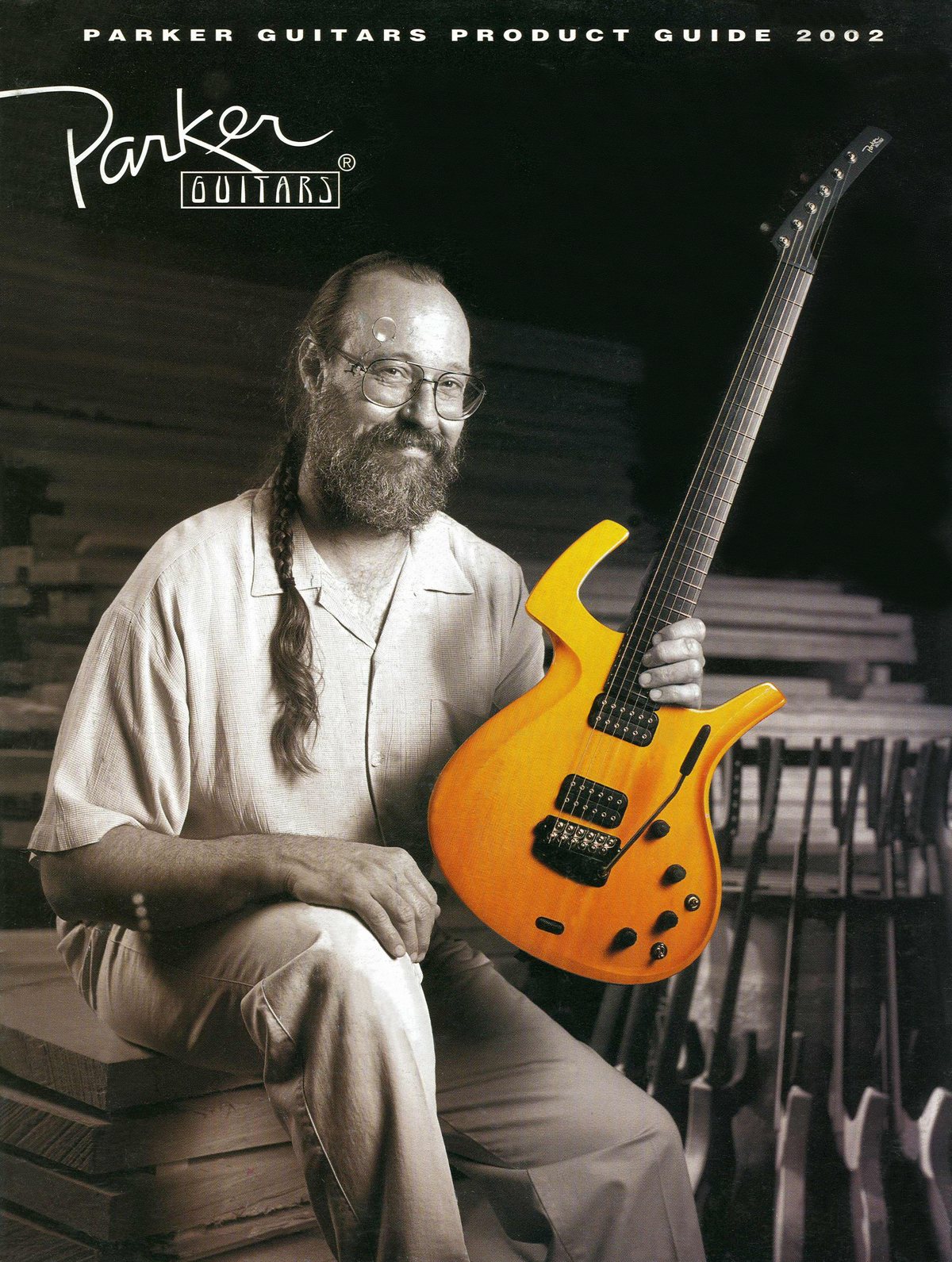 Cover of the Parker Guitars Product Guide 2002: a black and white photo of Ken Parker, bearded with a long braid and glasses, seated on stacked lumber in his workshop and holding an amber Parker Fly, the only thing in colour. Unfinished guitar necks stand in a rack behind him.