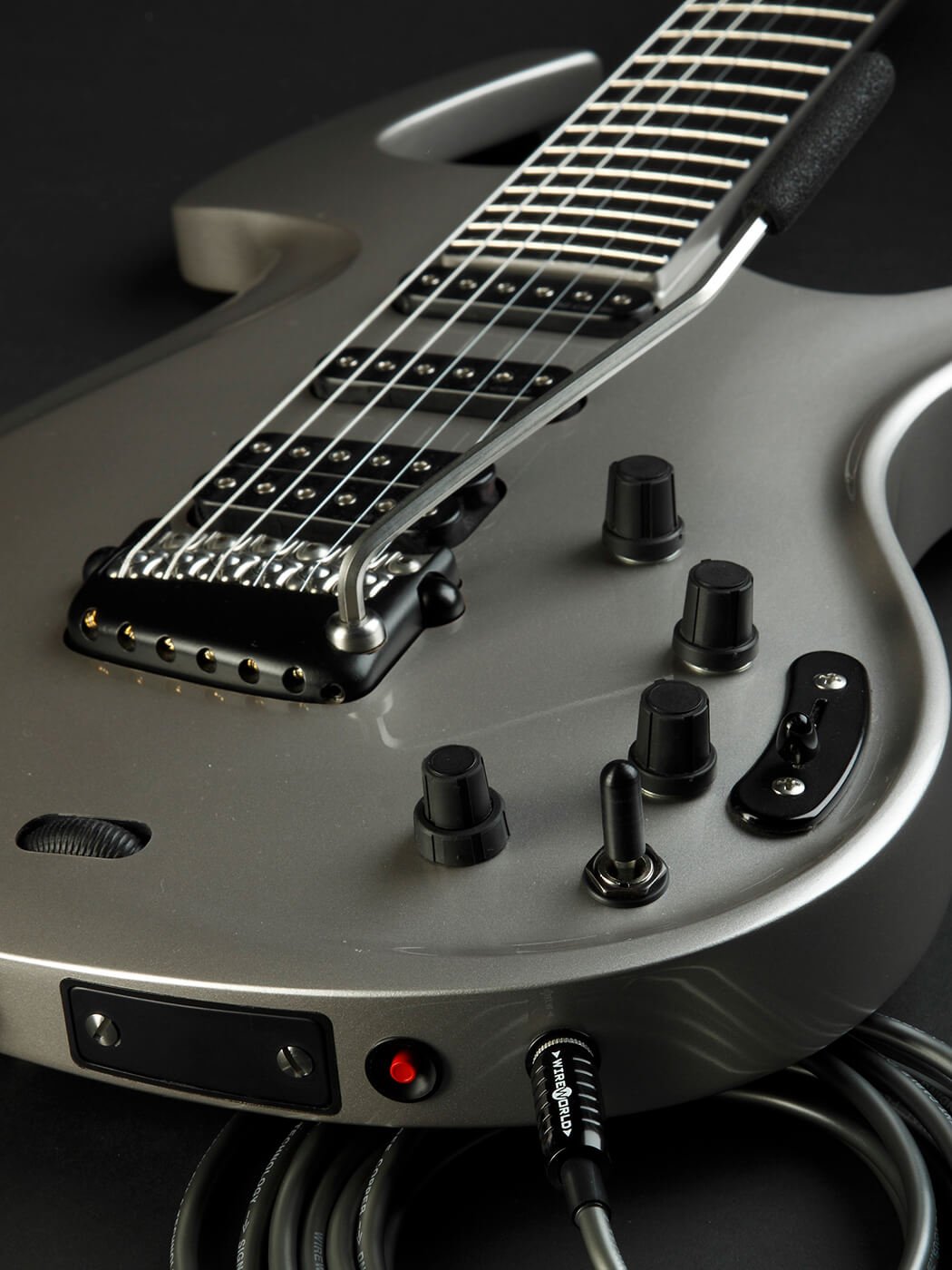 Close-up of a silver Parker Fly body on a black background: stainless steel frets, three pickups, the vibrato arm curving over the strings, four black control knobs and two toggle switches, with a cable plugged into the side.