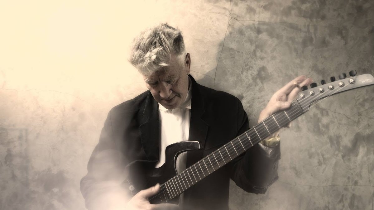 David Lynch, in a dark jacket and white shirt, leans against a cracked plaster wall and looks down at the Parker Fly he is playing, in warm sepia light.