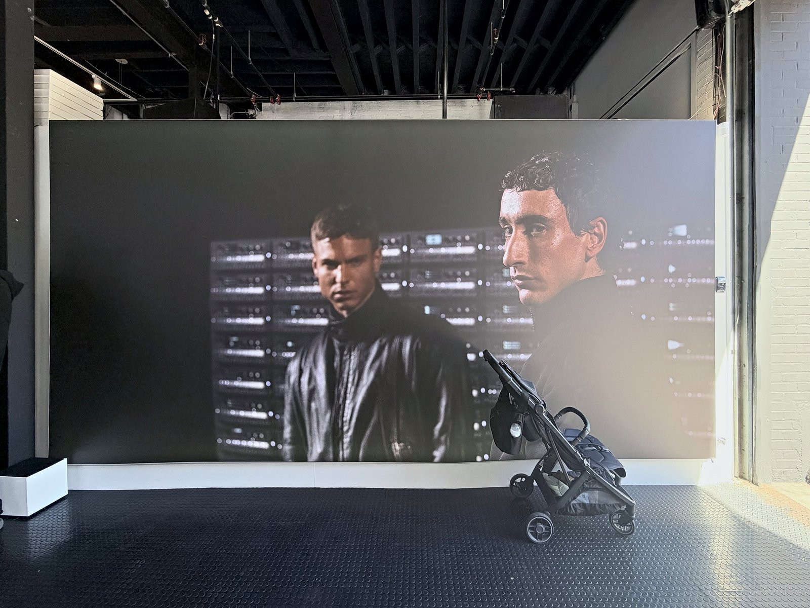 A large backlit print of two young men in dark jackets standing in front of stacked racks of glowing equipment, mounted on a wall of the store above a dark rubber floor. A folded stroller stands in front of it.