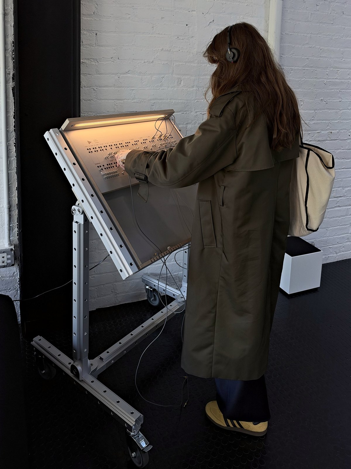 A visitor in headphones and a long olive coat patches cables into a large synthesizer panel mounted on a tilted aluminium stand on wheels.