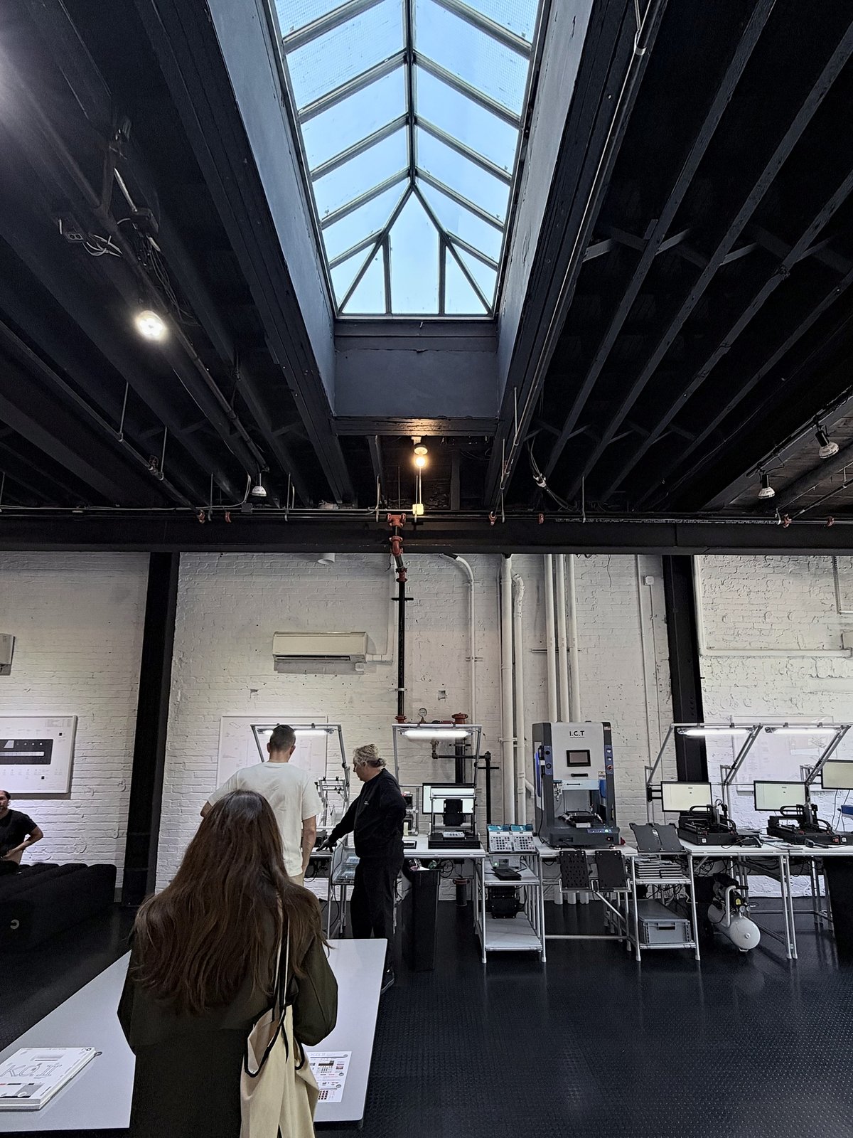 Visitors in a white-painted brick room under a long pitched skylight, watching staff at a row of workbenches with assembly and test machines, lamps and monitors.