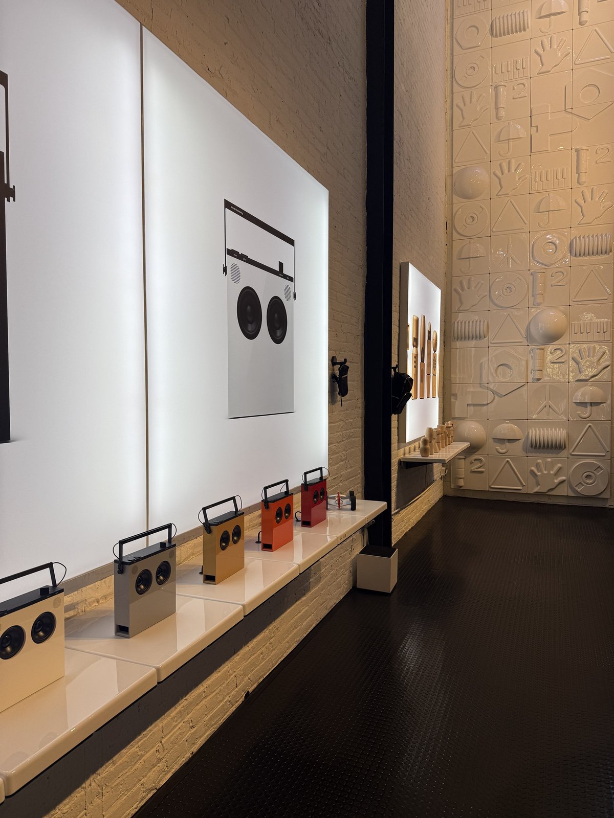 A row of OB-4 Magic Radios with carry handles in cream, grey, yellow, orange and red on a long white shelf, in front of lit panels showing a large speaker graphic. Beyond them, a wall of glazed white relief tiles with hands, umbrellas, triangles and discs.
