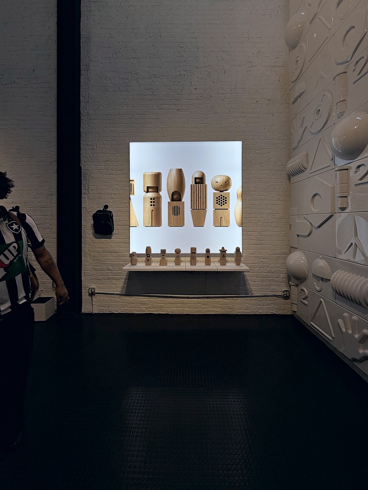 A lightbox showing a row of small wooden CH-8 Choir figures, each with a speaker or microphone grille, with the real figures lined up on a shelf beneath it against white-painted brick.